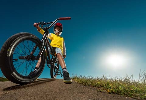 Photograph from a worm's eye view of a young boy on a bike, feet planted on the ground, wearing a helmet with a blue sky and sun in the background.